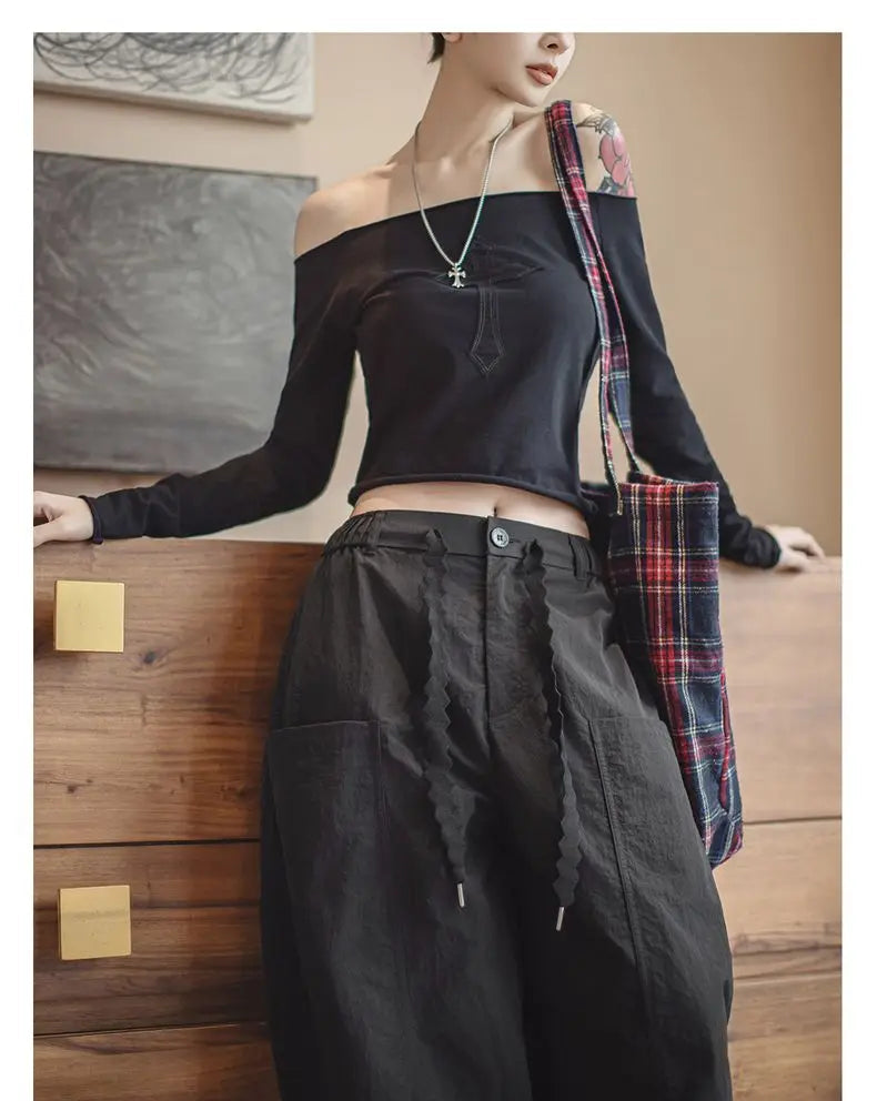 Person wearing black off-shoulder top and wide-leg pants with plaid bag, standing against a wooden wall.